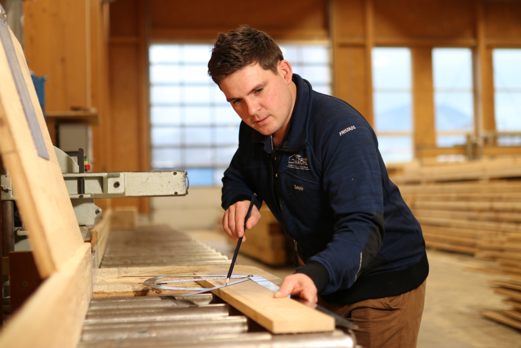 Ein Handwerker misst ein Holzbrett in einer hellen Werkhalle.